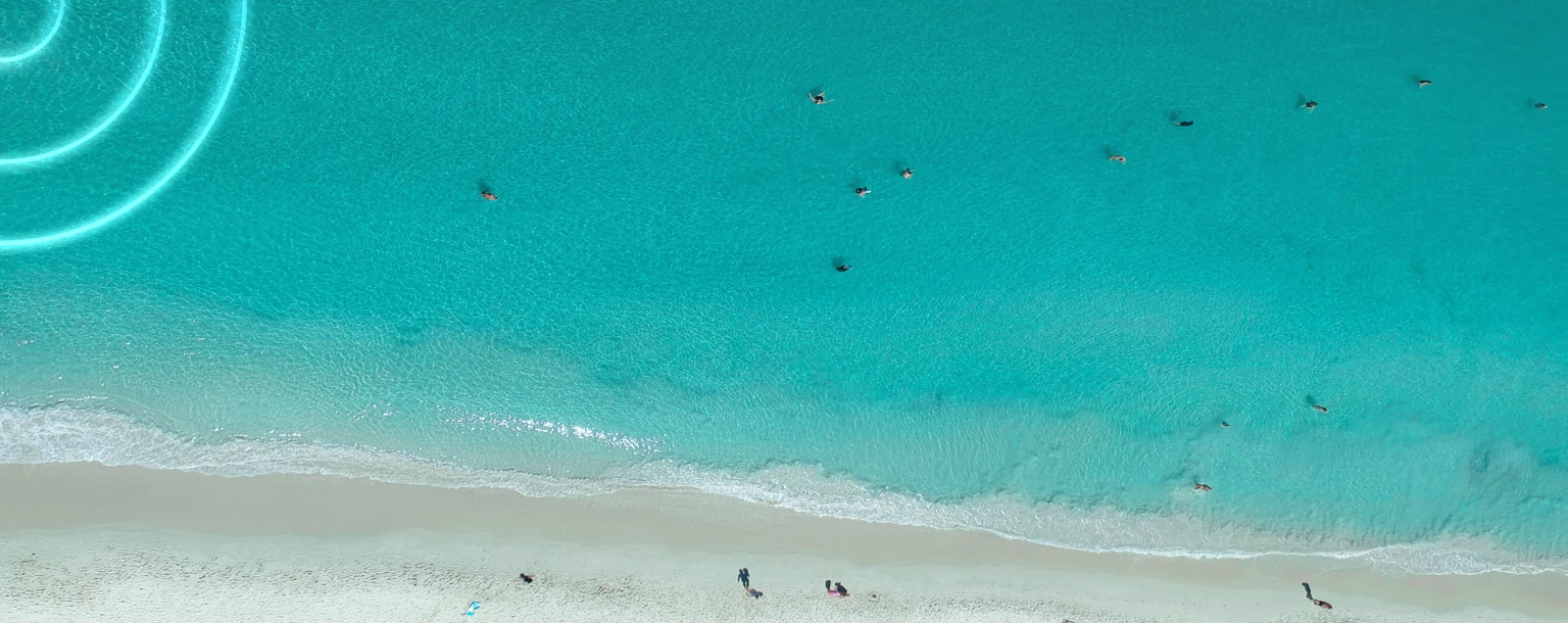 Aerial view of a turquoise beach with people swimming in clear water and sunbathers relaxing on the sandy shore.