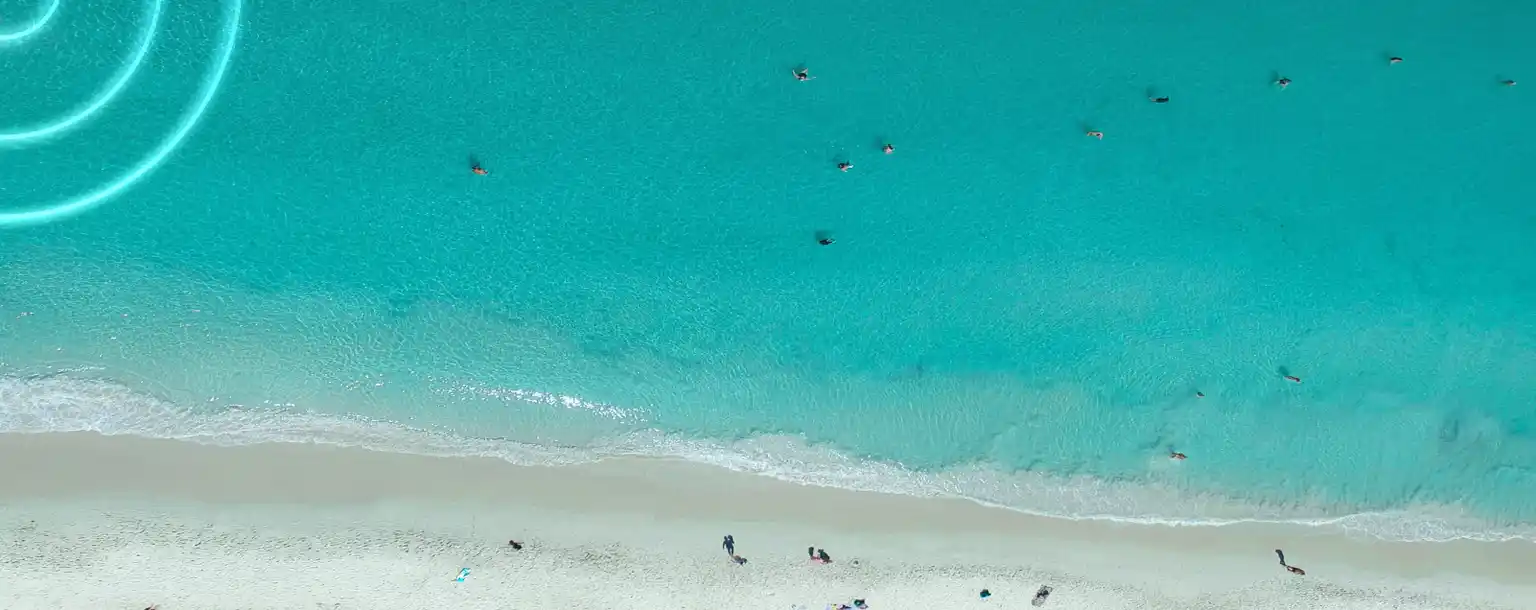 Aerial view of a turquoise beach with people swimming in clear water and sunbathers relaxing on the sandy shore.