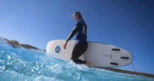 A surfer in a wetsuit carries a surfboard through shallow, clear blue water under a bright blue sky.