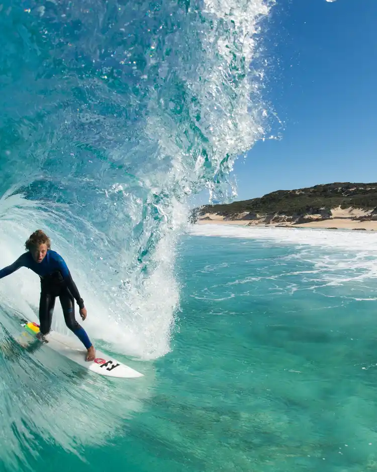Surfer riding inside a clear blue wave tube near a sandy beach under a sunny sky.