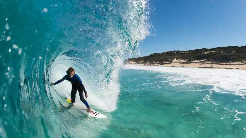 Surfer riding inside a clear blue wave tube near a sandy beach under a sunny sky.