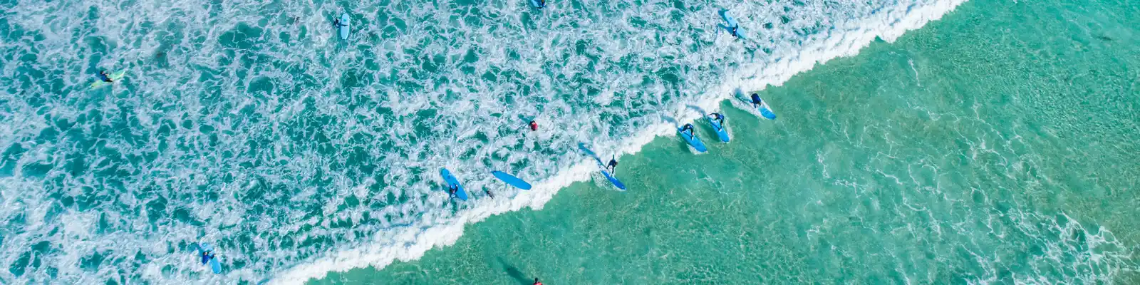Aerial view of surfers paddling on turquoise waves near the shore, creating white surf patterns and vibrant splashes in the ocean.