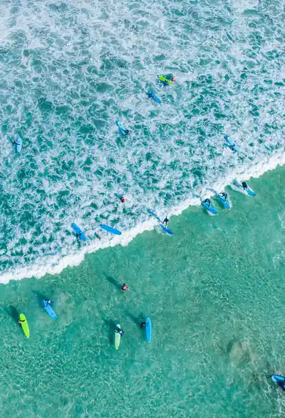 Aerial view of surfers paddling on turquoise waves near the shore, creating white surf patterns and vibrant splashes in the ocean.