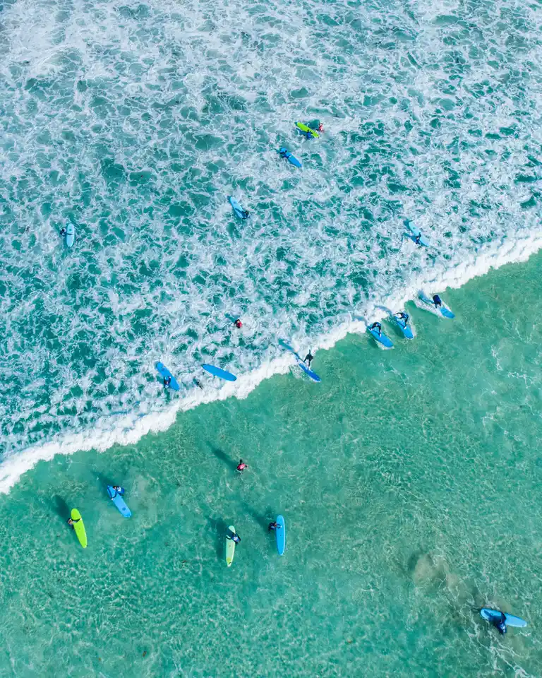 Aerial view of surfers paddling on turquoise waves near the shore, creating white surf patterns and vibrant splashes in the ocean.