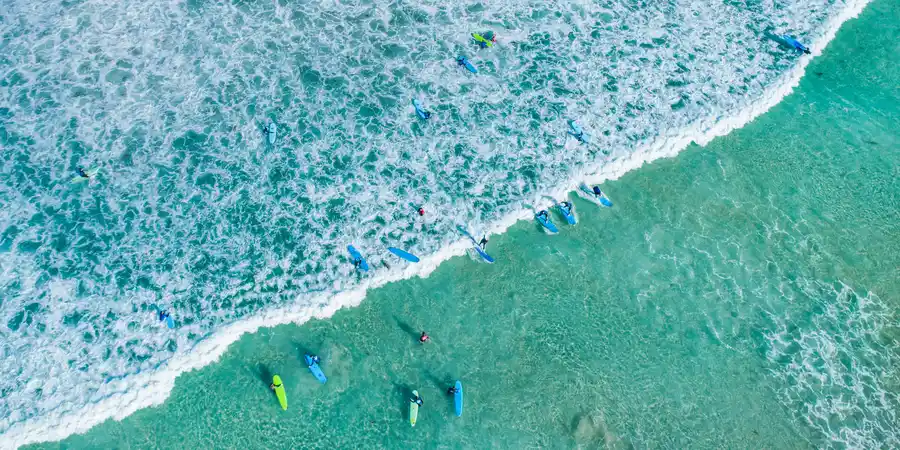 Aerial view of surfers paddling on turquoise waves near the shore, creating white surf patterns and vibrant splashes in the ocean.