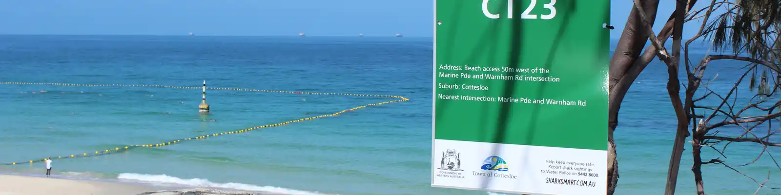 Beach scene with a sign reading "Cottesloe Beach CT23," clear blue sea, yellow buoys, and trees in the foreground.