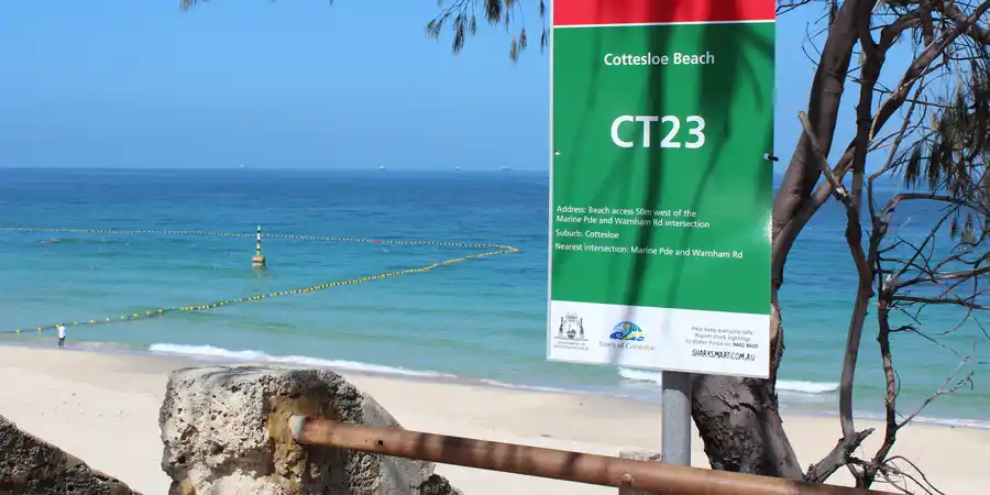 Beach scene with a sign reading "Cottesloe Beach CT23," clear blue sea, yellow buoys, and trees in the foreground.
