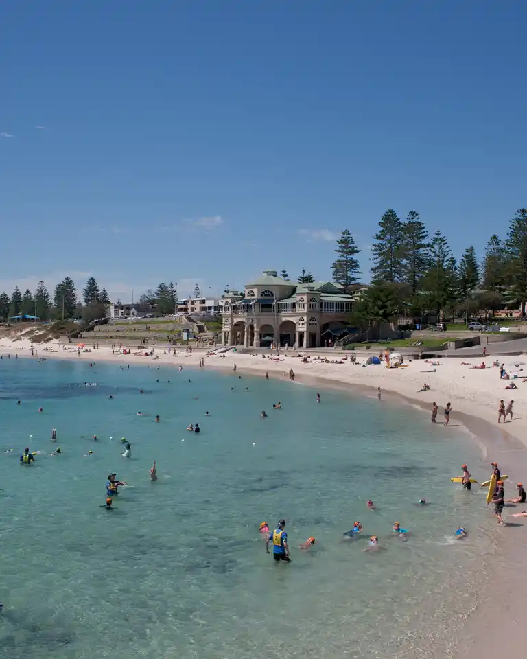 Sunny beach scene with people swimming and relaxing on the sandy shore, lined with trees and buildings in the background.