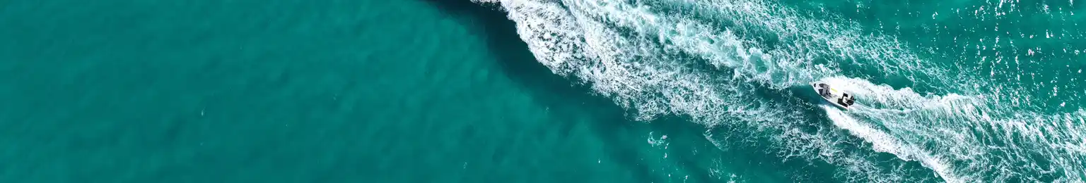 Aerial view of a boat cruising through clear turquoise waters, leaving a trail of white wake, with gentle waves in the background.