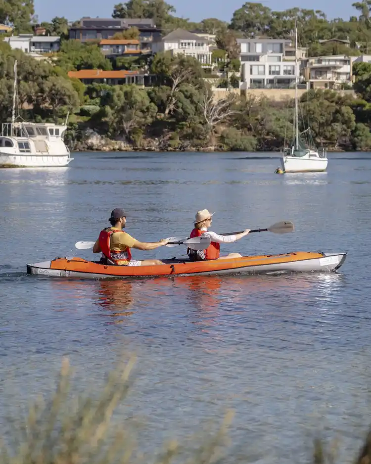 Two people kayaking in an orange kayak on a calm lake, with boats and houses on a tree-lined shore in the background.