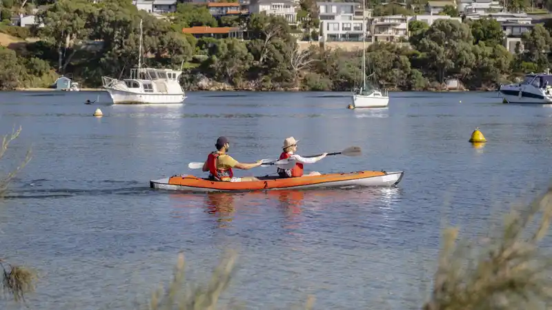 Two people kayaking in an orange kayak on a calm lake, with boats and houses on a tree-lined shore in the background.