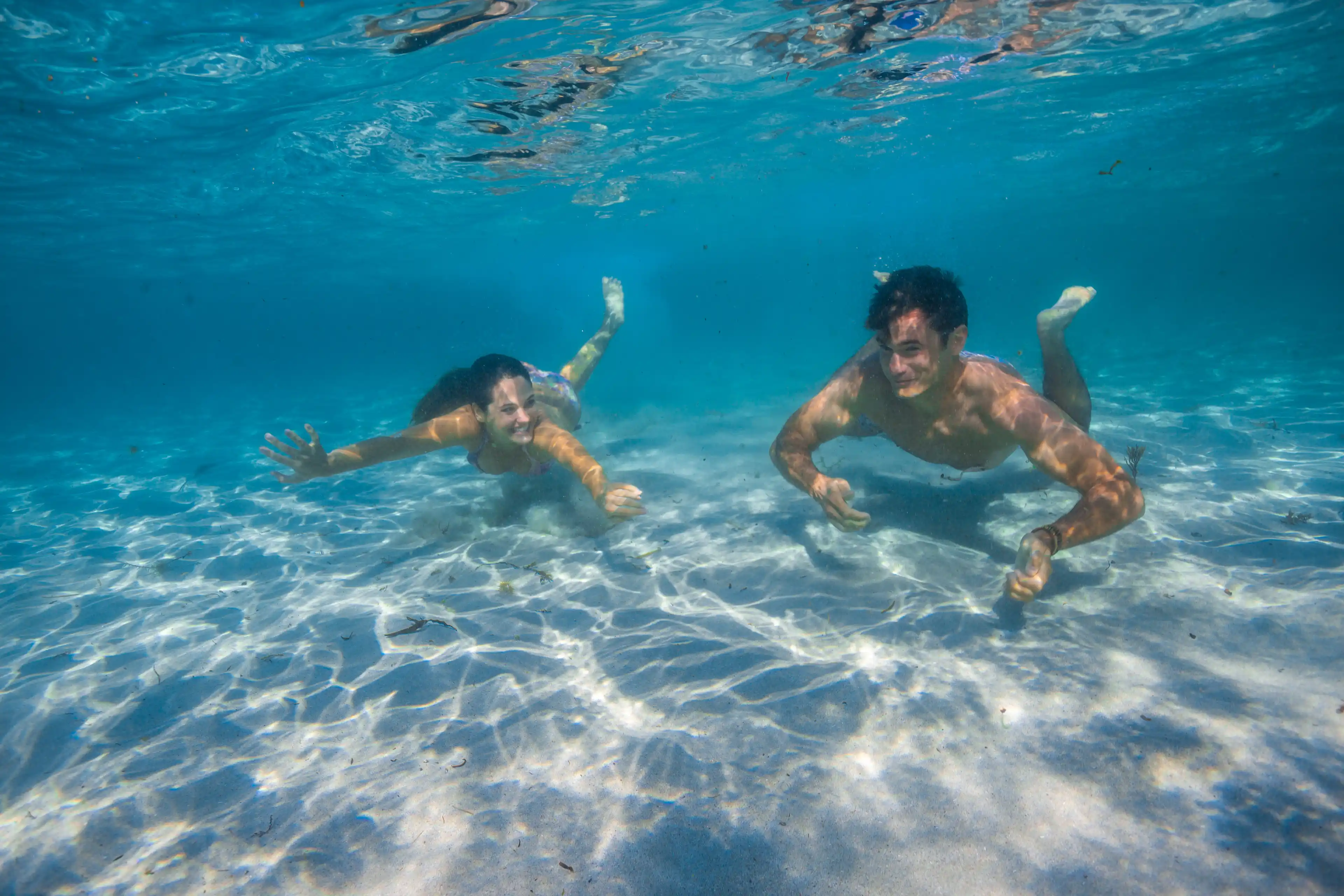 Two people swimming underwater in clear blue sea, sunlight creating ripples on the sandy bottom.