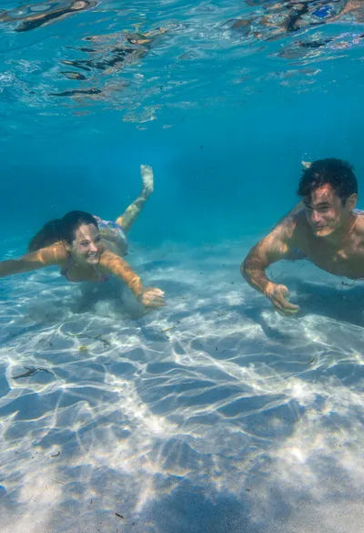 Two people swimming underwater in clear blue sea, sunlight creating ripples on the sandy bottom.
