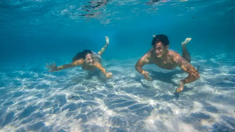 Two people swimming underwater in clear blue sea, sunlight creating ripples on the sandy bottom.