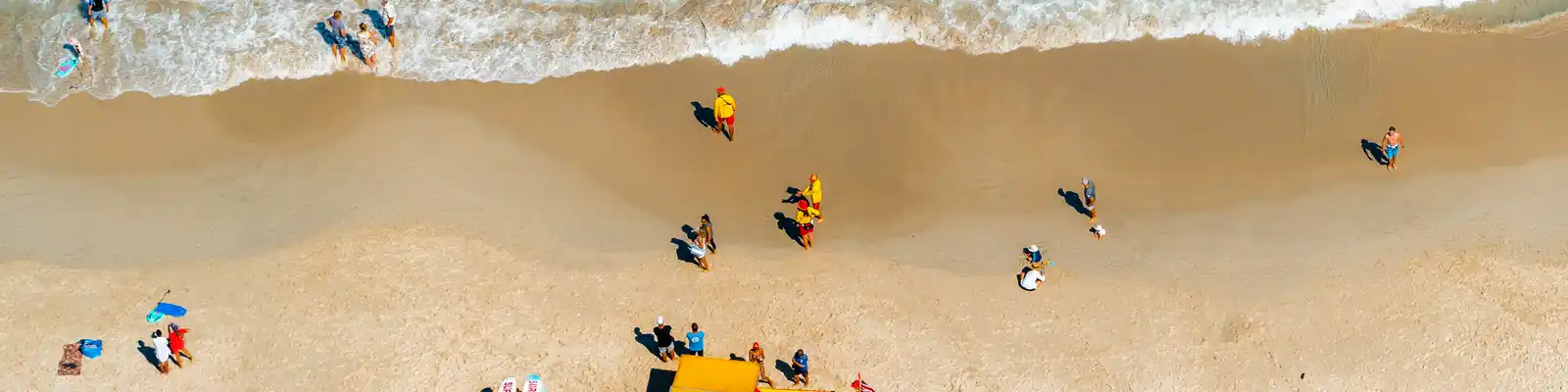 Aerial view of a sandy beach with people sunbathing, umbrellas, surfboards, and a lifeguard vehicle near turquoise waves.