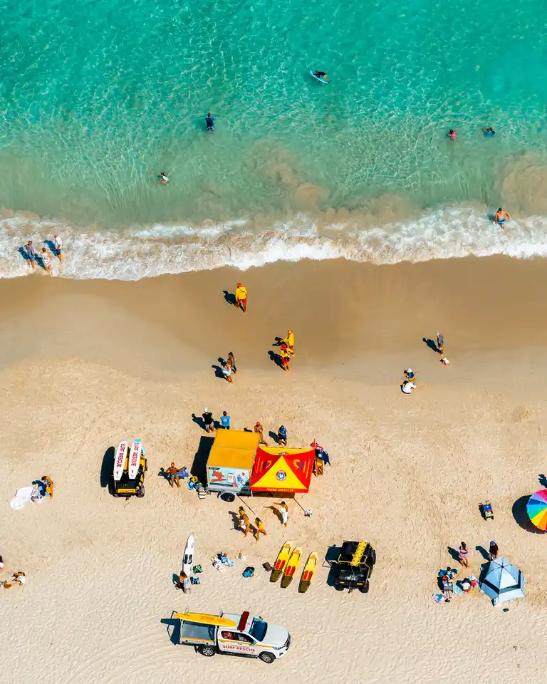 Aerial view of a sandy beach with people sunbathing, umbrellas, surfboards, and a lifeguard vehicle near turquoise waves.