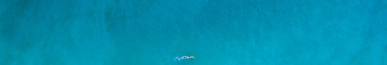 Aerial view of four swimmers in clear turquoise ocean water near a sandy beach with gentle waves.