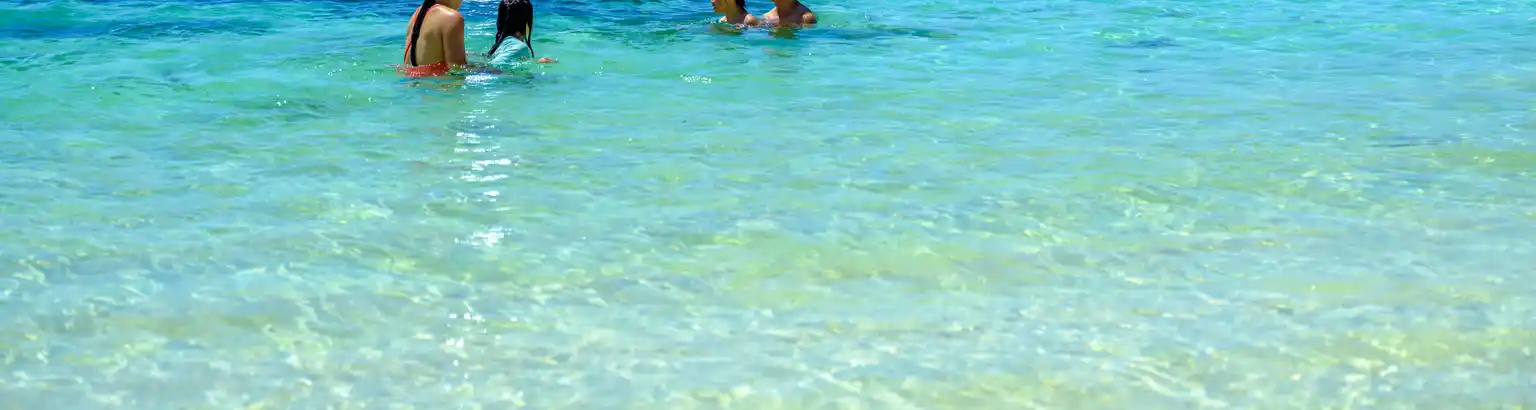 A family enjoys swimming in clear turquoise waters with boats anchored in the distance under a bright blue sky.