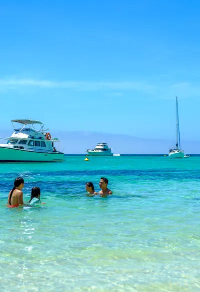 A family enjoys swimming in clear turquoise waters with boats anchored in the distance under a bright blue sky.