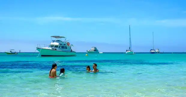 A family enjoys swimming in clear turquoise waters with boats anchored in the distance under a bright blue sky.