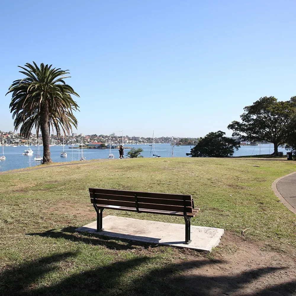 A park with a bench facing a bay, palm tree nearby, and boats on the water. Clear blue sky and trees along the shoreline.