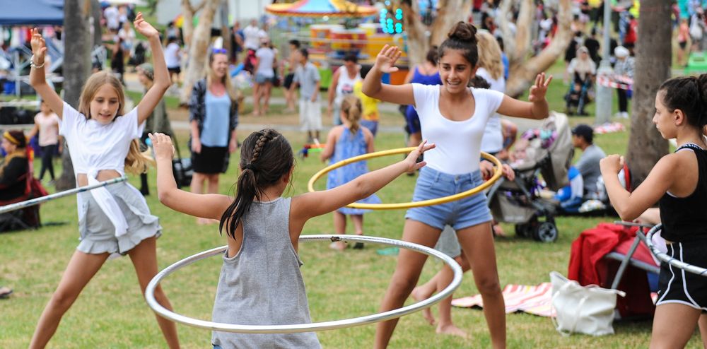 Children playing with hula hoops in a park during a lively outdoor event, with trees, people, and amusement rides in the background.