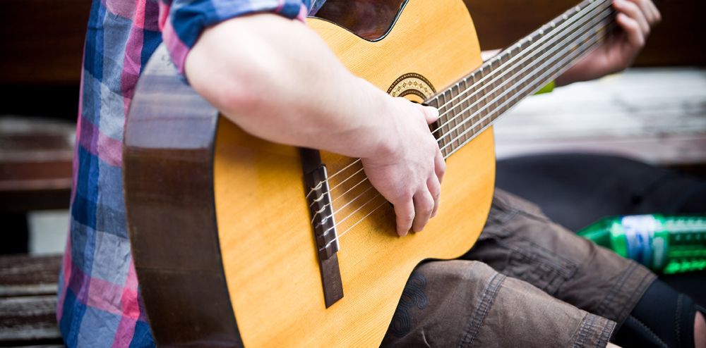 Person playing an acoustic guitar, wearing a plaid shirt and shorts, sitting outdoors with a green bottle nearby.