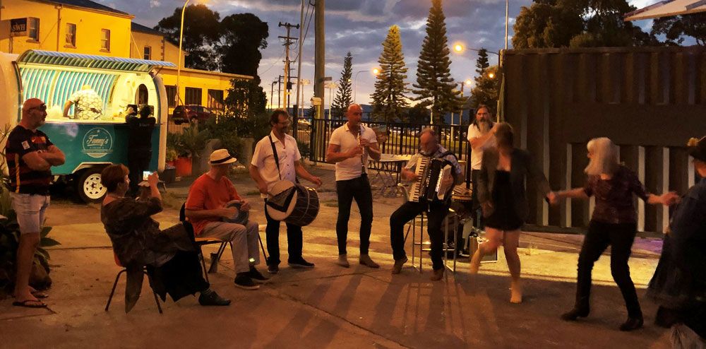 People enjoying live music outdoors in the evening, with musicians playing accordion and drums, and others dancing near a food truck.