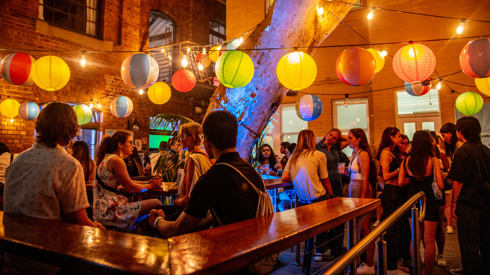 People socializing at an outdoor party with colorful lanterns and warm lighting, creating a festive atmosphere against a brick building backdrop.