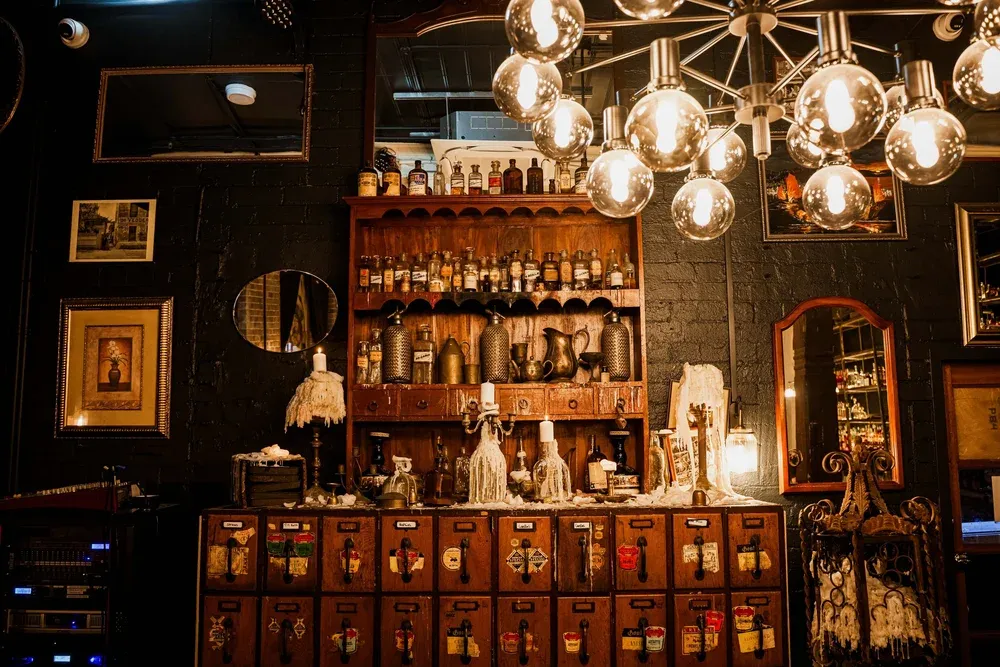 Vintage apothecary setting with wooden cabinets, labeled drawers, antique bottles, and a modern chandelier illuminating the space.