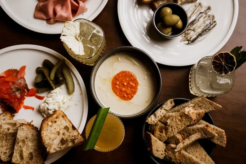A table with plates of assorted appetizers: cured meats, pickles, bread, olives, anchovies, and dip. Two cocktails are also visible.