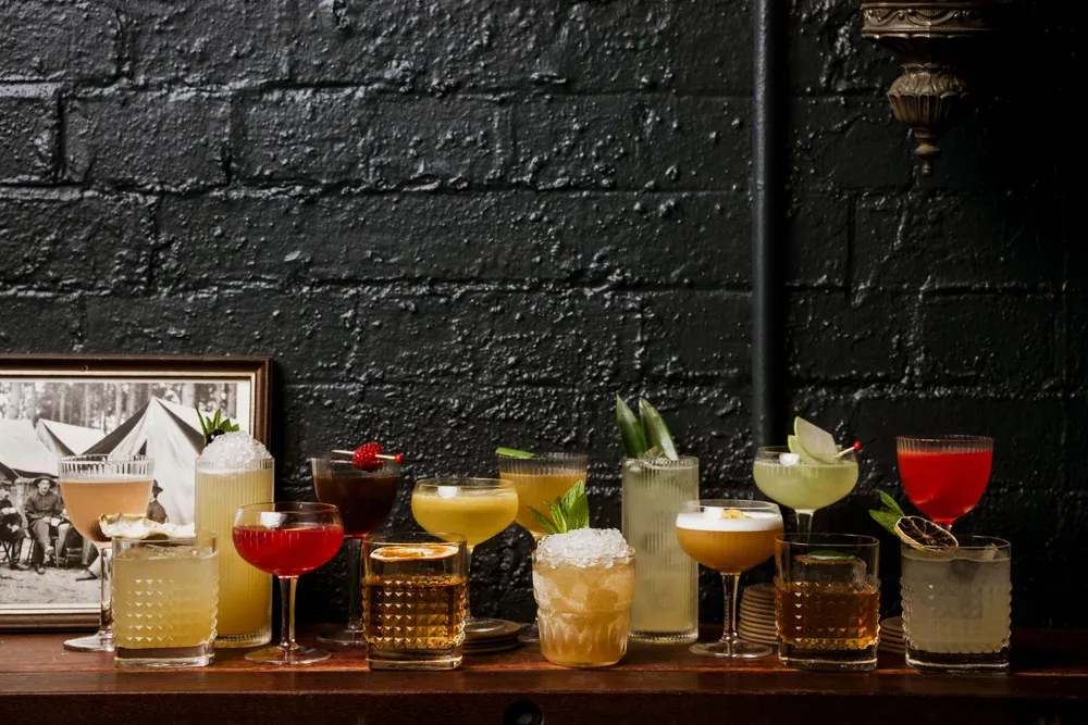A variety of colorful cocktails in different glasses arranged on a wooden surface against a textured black brick wall. A framed photo is on the left.