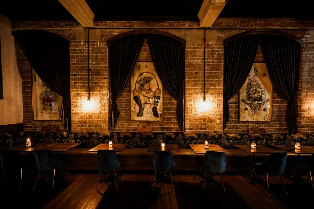 A dimly lit, cosy bar interior with brick walls, dark drapes, framed art, and candles on tables, creating an intimate atmosphere.