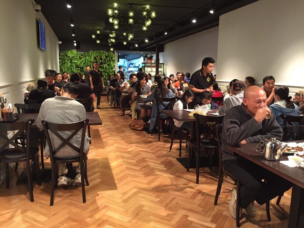 A busy restaurant with patrons dining at tables, waitstaff serving, and a green plant wall in the background.