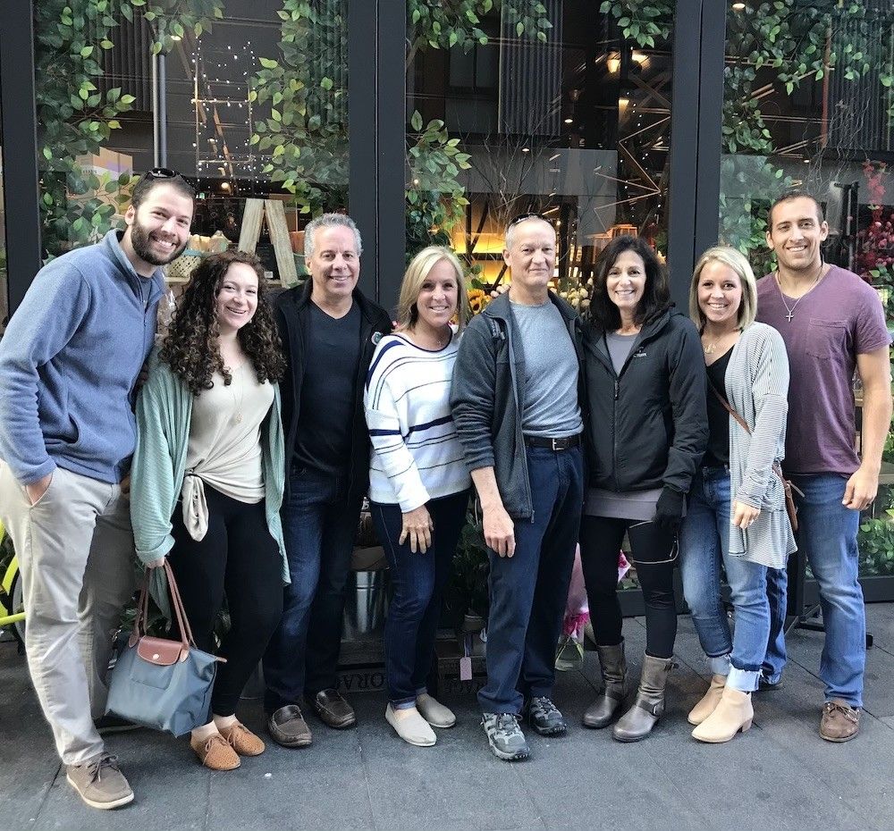 A group of eight people smiling and standing in front of a building with greenery.