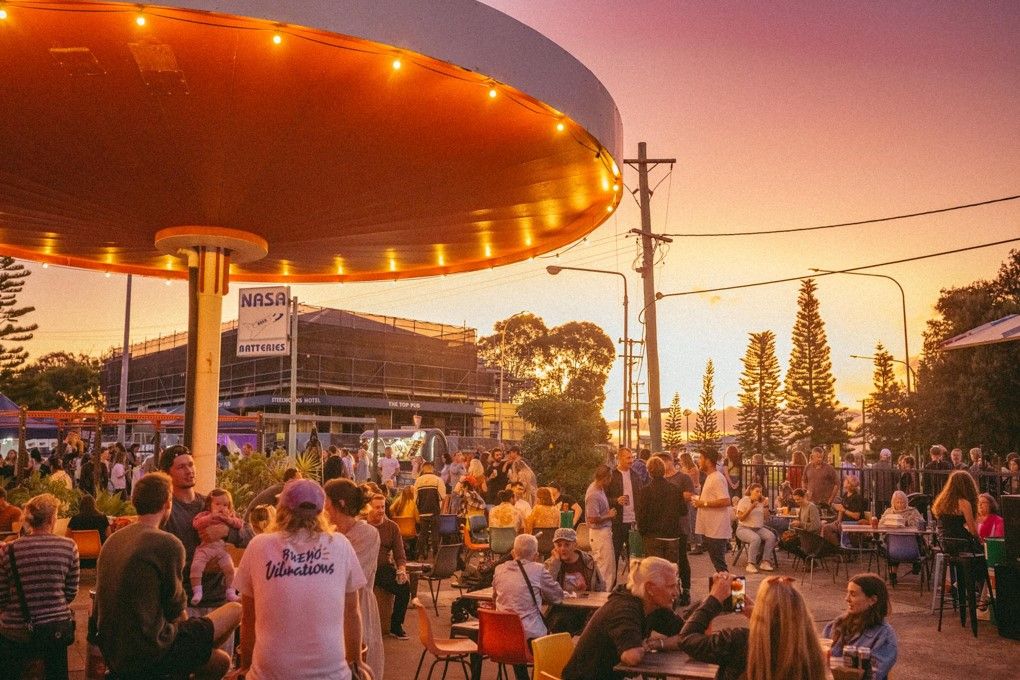 Outdoor gathering at sunset with people sitting and standing under a round structure with string lights, set against a pink and orange sky.