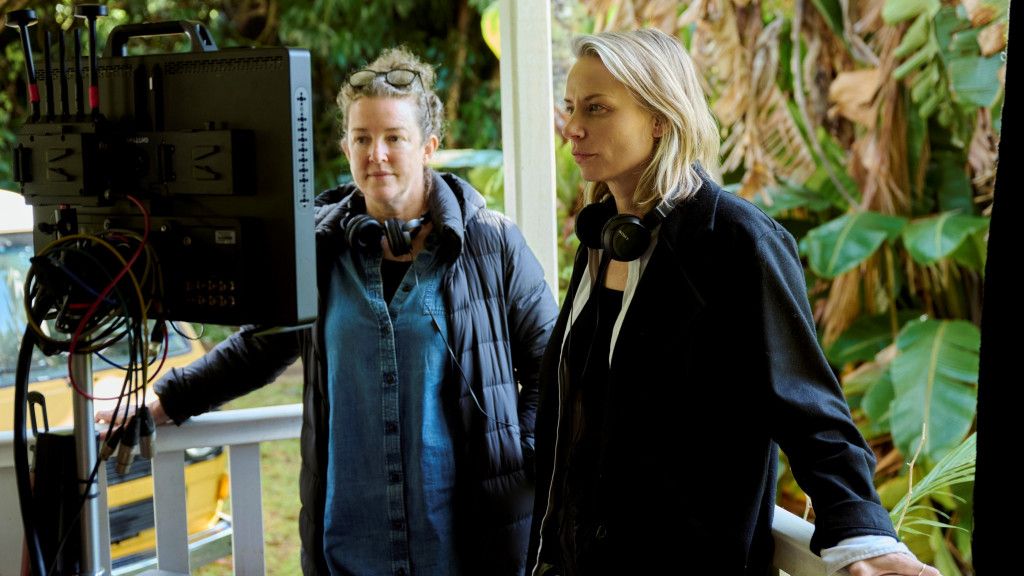 Two people stand outdoors, intently watching a monitor, wearing headphones, with lush greenery in the background.