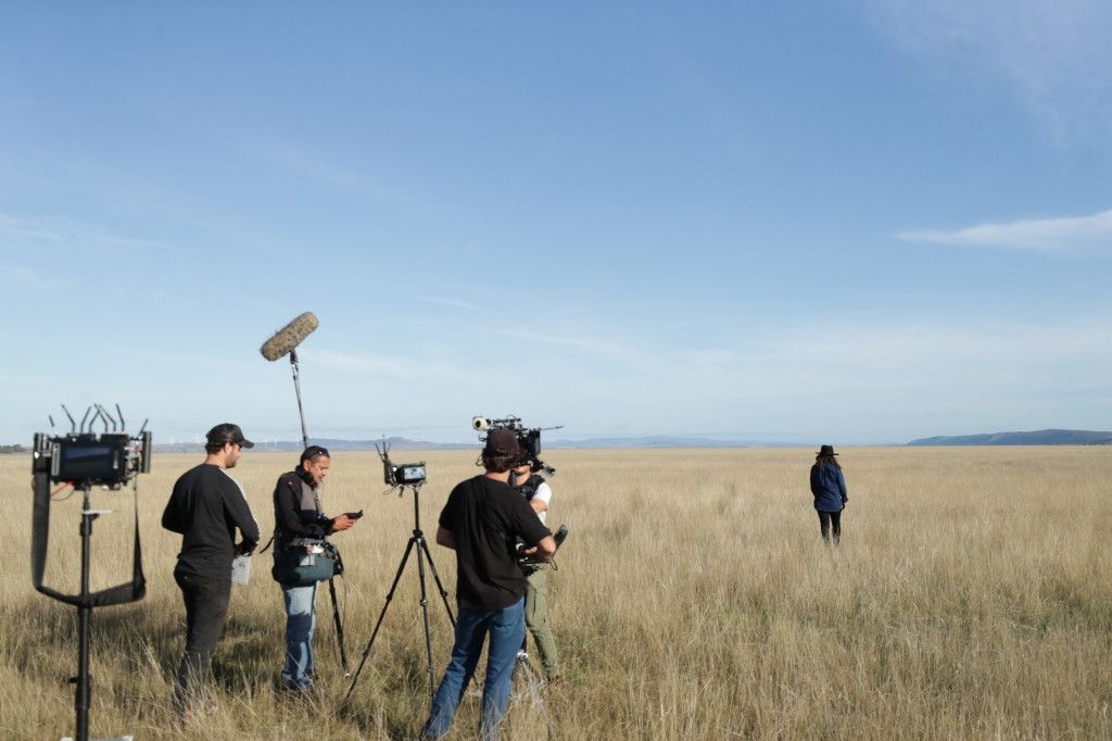 Film crew with cameras and microphones set up in an open field, capturing a scene of a person walking away in the distance.