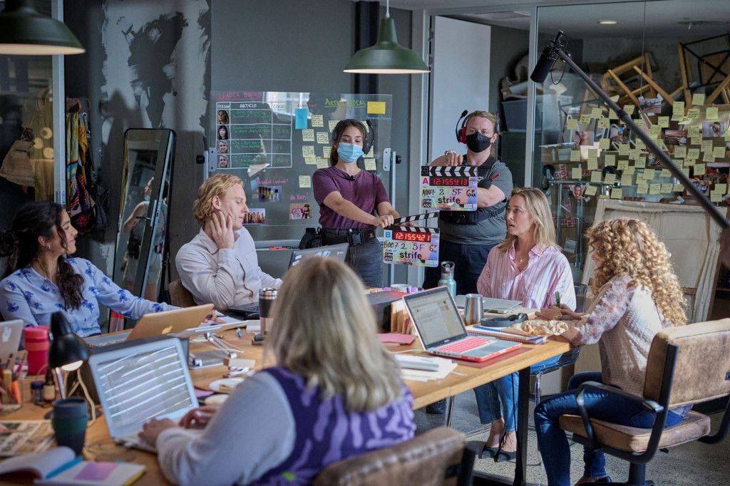 A group of people in a meeting room discuss around a table with laptops, while two crew members prepare a scene with a clapperboard.
