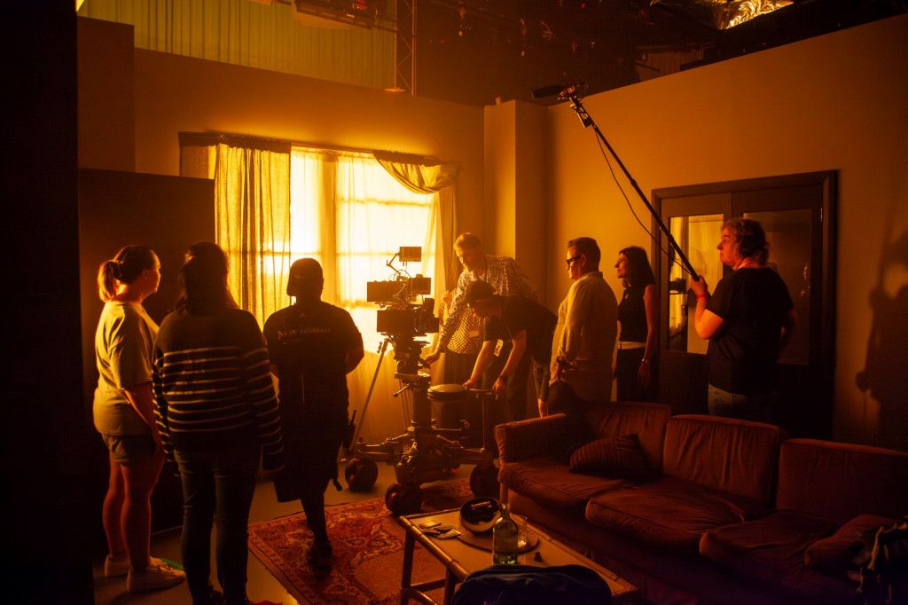 Film crew setting up in a dimly lit room with a brown couch, camera, and boom mic next to a window casting warm light inside.
