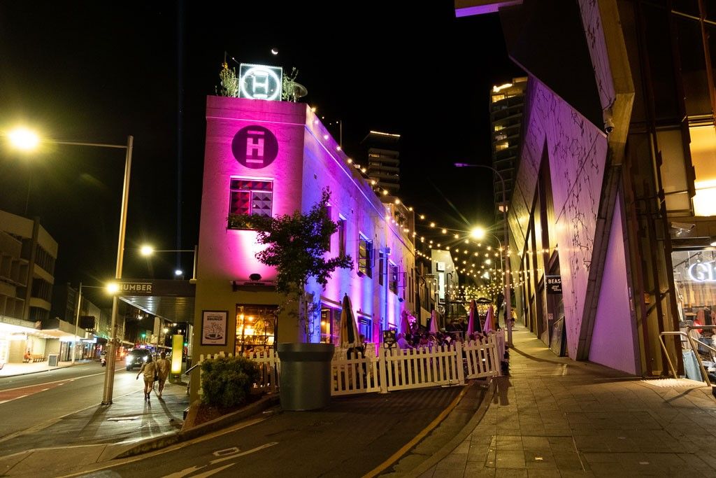 Nighttime street scene with a building illuminated in pink and purple lights, surrounded by string lights and a small outdoor seating area.