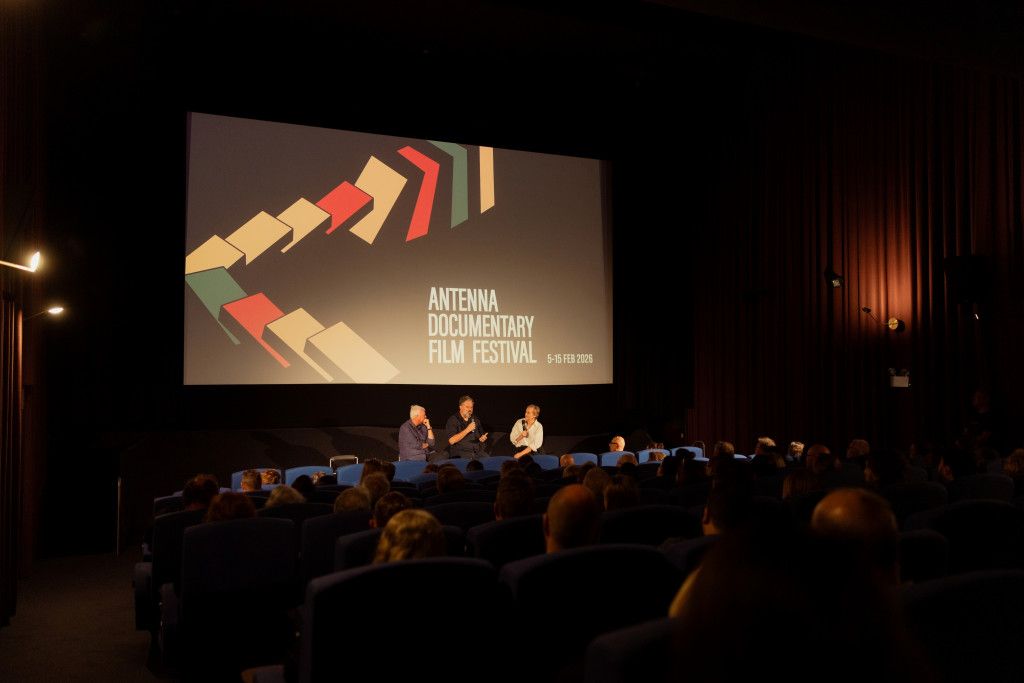 People in a dimly lit cinema attend the Antenna Documentary Film Festival, with a large screen displaying the festival's logo.