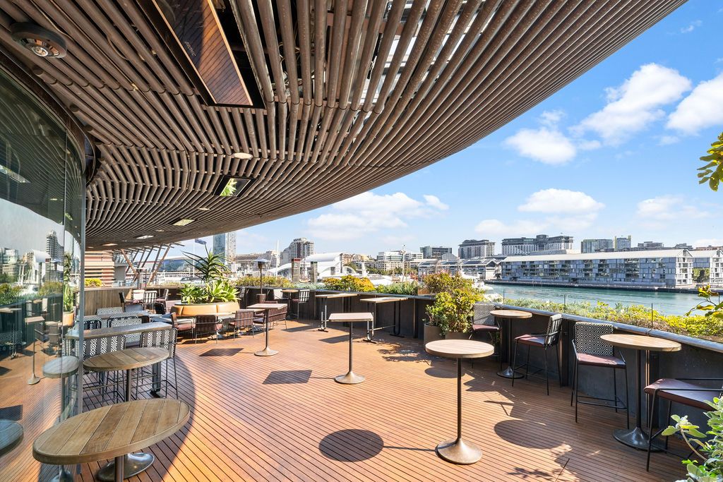 Modern rooftop terrace with wooden flooring, tables, and chairs overlooking a river and city skyline under a clear blue sky.