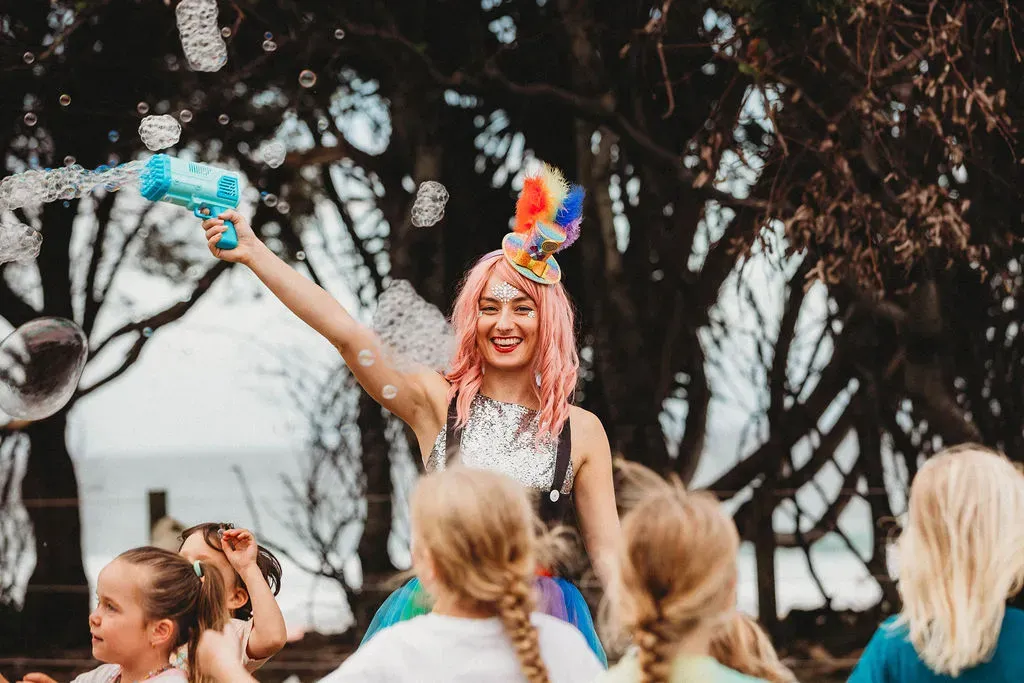 Performer in a colourful outfit and pink wig entertains children by blowing bubbles with a toy gun in an outdoor setting.