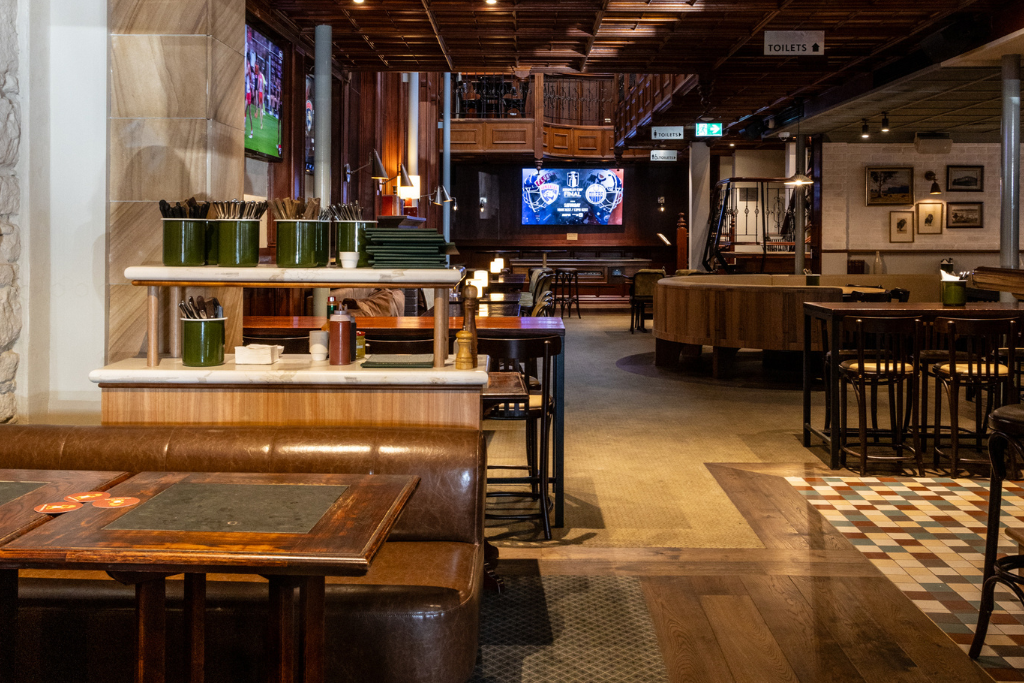 Cozy pub interior with wooden tables, chairs, and a shelf for condiments. Flat-screen TV displays a sports game. Warm lighting and tiled floor.