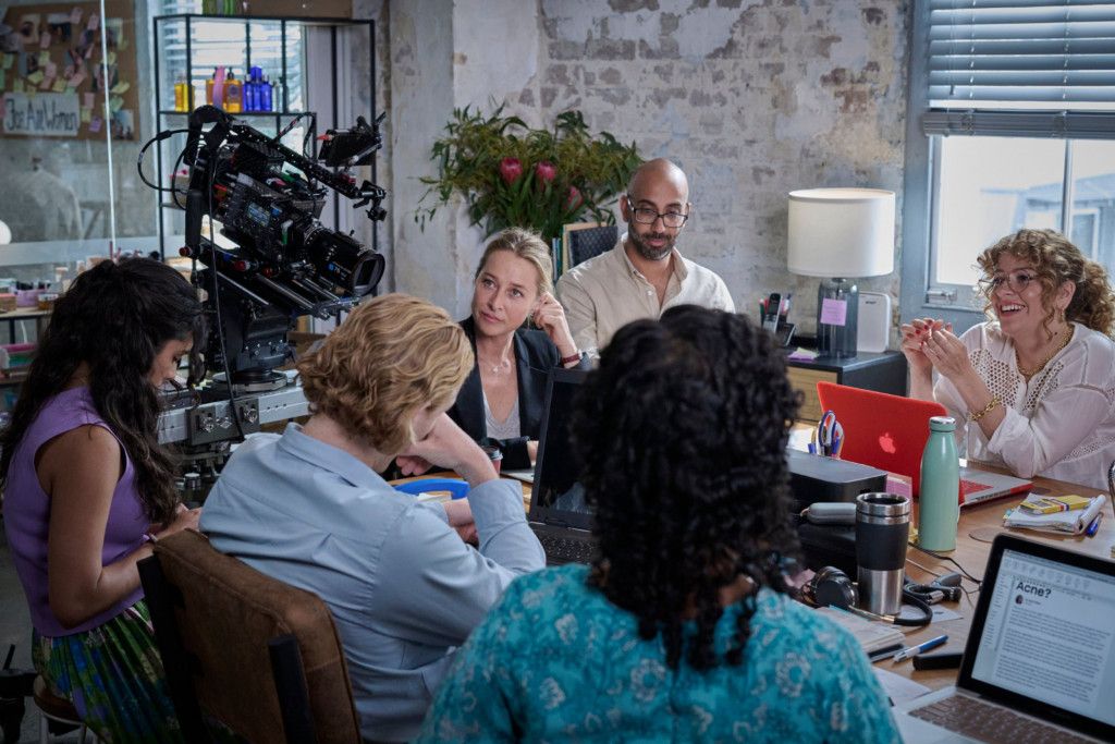 A group of people sit around a table with laptops and papers, engaged in a discussion. A camera is set up nearby in an industrial-style room.