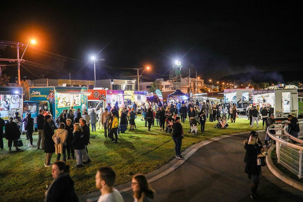 A nighttime food truck festival with crowds of people gathered around brightly lit trucks on a grassy area.