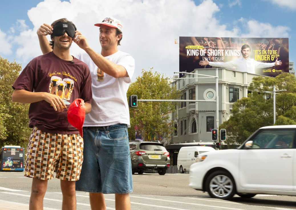 two people one with eye mask on standing in front of billboard at taylor square