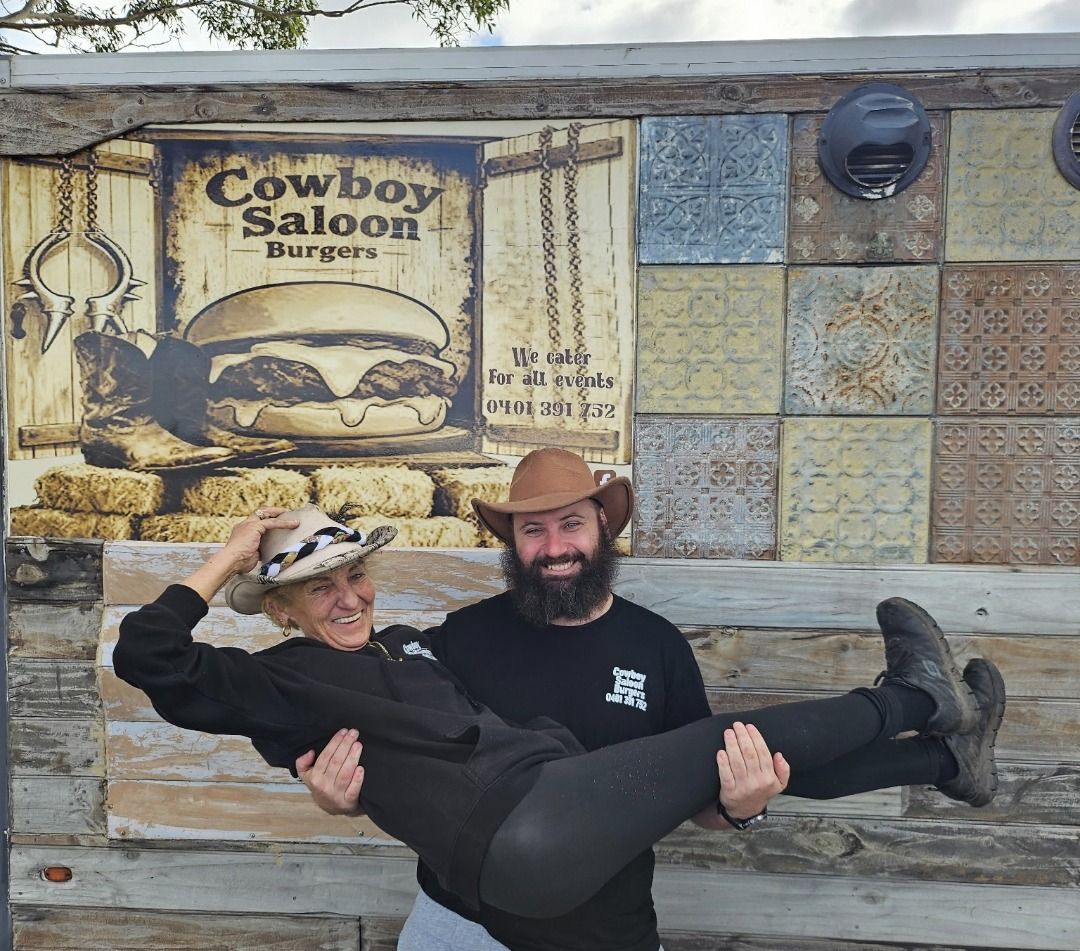 Two people wearing cowboy hats, one carrying the other, stand in front of a "Cowboy Saloon Burgers" sign on a rustic wall.