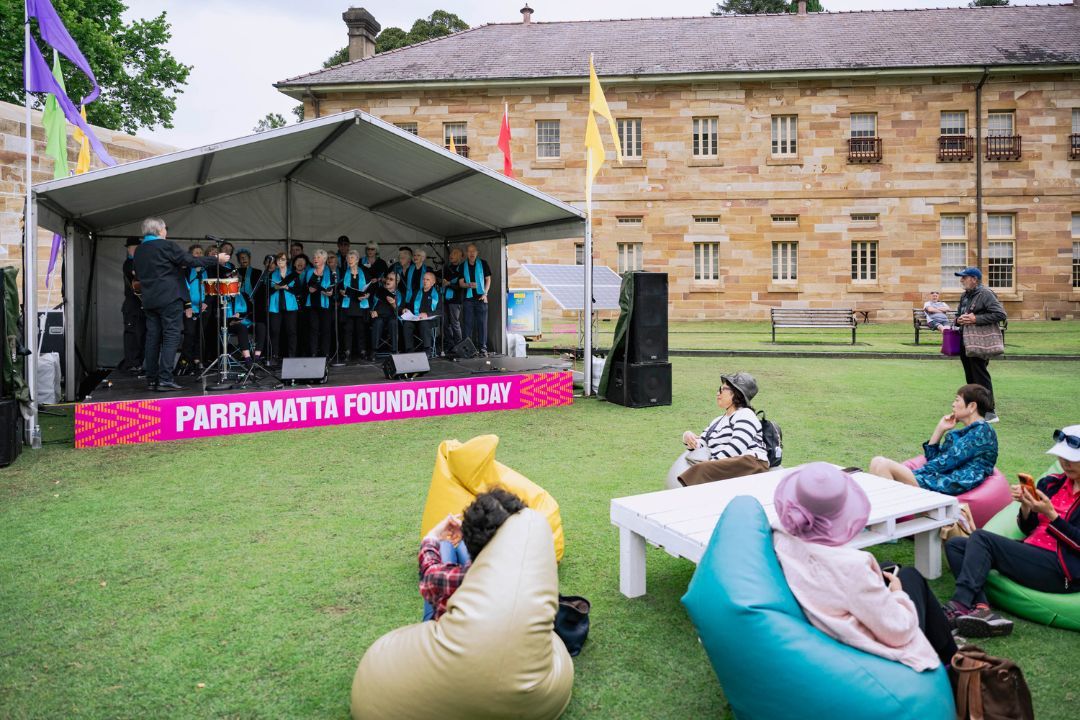 A stage that has the words Foundation Day across the bottom, with a large group pf people in blue on the stage. Some people are seated in front of the stage watching the performers.